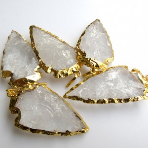 May include: Five clear quartz arrowhead pendants with gold-plated edges. The pendants are arranged in a cluster on a white background.