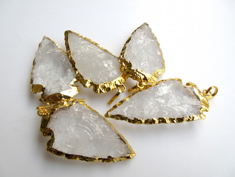 May include: Five clear quartz arrowhead pendants with gold-plated edges. The pendants are arranged in a cluster on a white background.