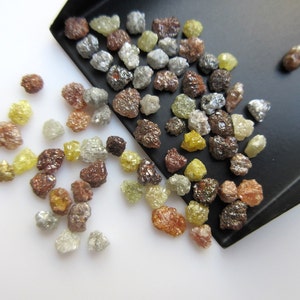 May include: A collection of rough, uncut diamonds in various colors, including brown, gray, yellow, and clear. The diamonds are scattered on a white surface and in a black tray.