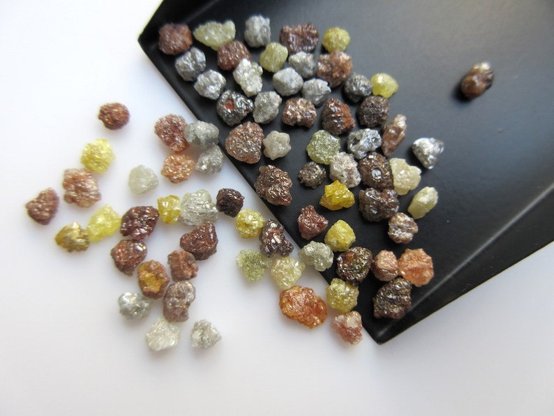 May include: A collection of rough, uncut diamonds in various colors, including brown, gray, yellow, and clear. The diamonds are scattered on a white surface and in a black tray.