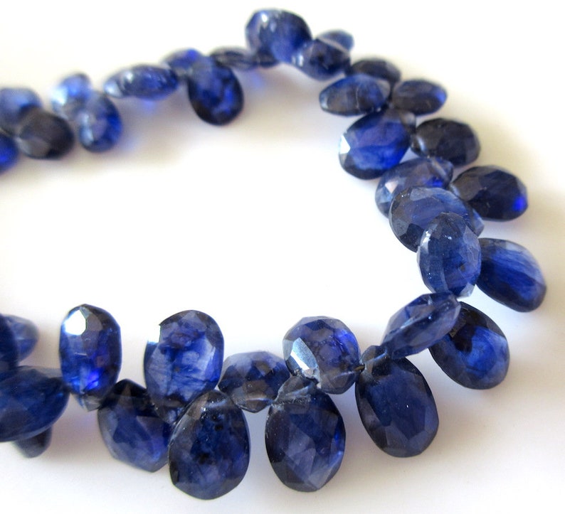 May include: A strand of dark blue gemstone beads, each bead is faceted and oval shaped. The beads are strung together to form a necklace or bracelet.