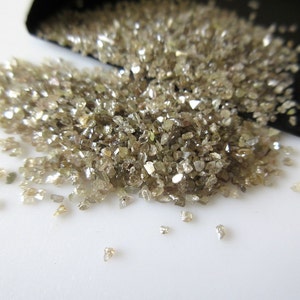 May include: A pile of small, rough, brown diamonds, some with a slight sparkle, spilling out of a black container onto a white surface.