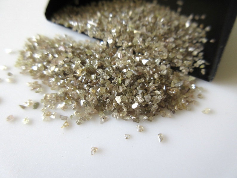 May include: A pile of small, rough, brown diamonds, some with a slight sparkle, spilling out of a black container onto a white surface.
