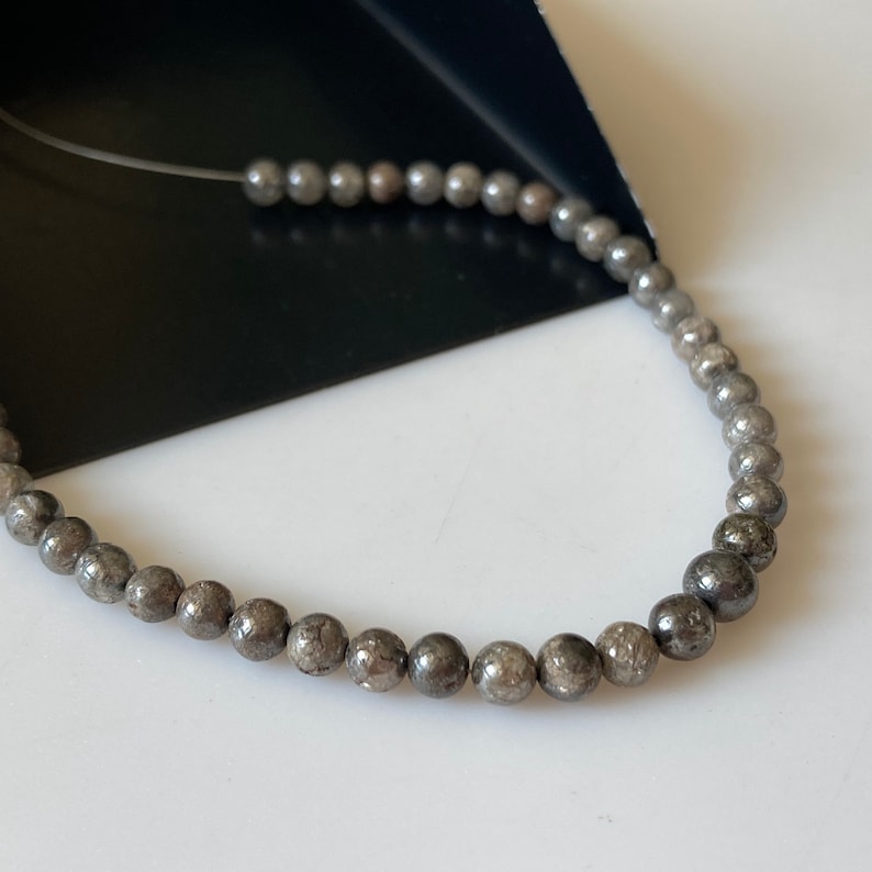 May include: A strand of small, round, gray beads. The beads are strung on a thin, clear cord. The beads have a metallic sheen and are evenly spaced along the cord. The background is a mix of white and black.