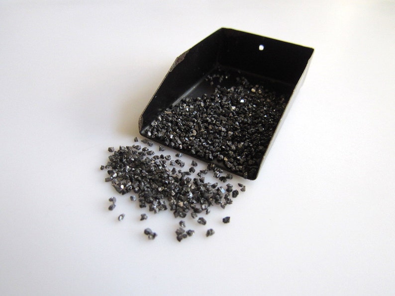 May include: A black metal scoop filled with small, dark gray, irregularly shaped pieces of material. Some of the material has spilled out of the scoop onto a white surface.