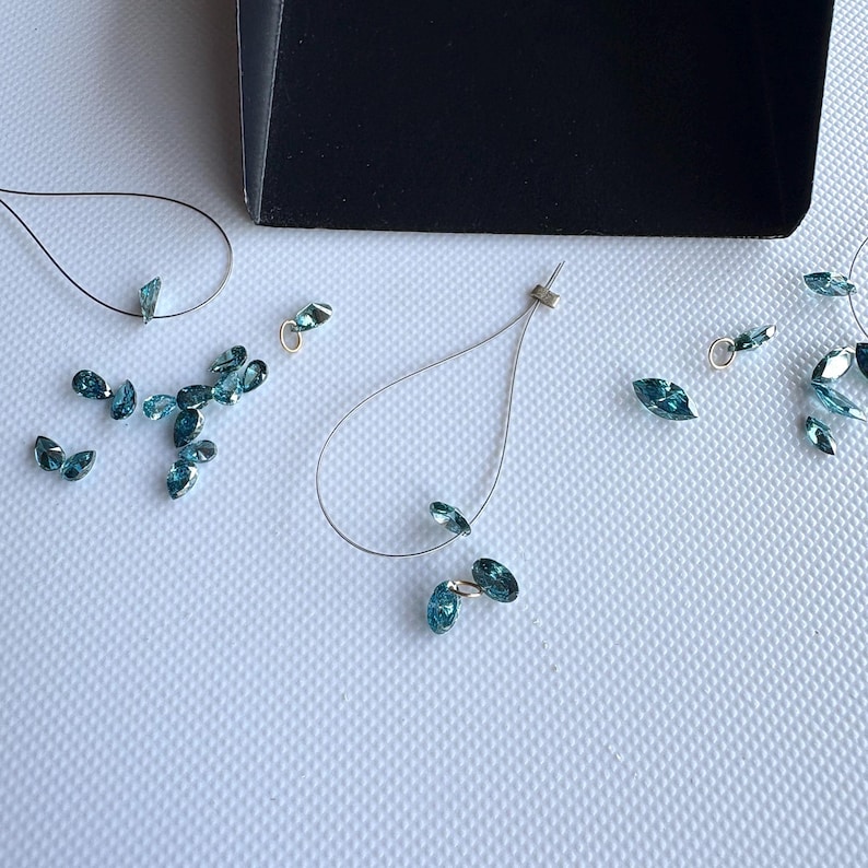 May include: Teardrop-shaped blue-green crystal beads and wire jewelry components scattered on a white surface. A black box is in the background. The crystals are in various sizes and shapes, suggesting a craft or jewelry-making theme.