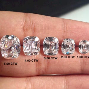 May include: Five cushion-cut diamonds of varying sizes are displayed, ranging from 1.00 CTW to 5.00 CTW. The diamonds are clear and faceted, reflecting light. The diamonds are set against a neutral background.