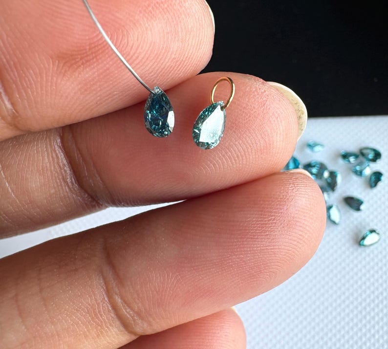 May include: Two small, pear-shaped, teal-colored gemstones. One gemstone is threaded on a thin silver wire, and the other is attached to a small gold-toned metal jump ring. Several additional loose teal pear-shaped gemstones are scattered nearby.