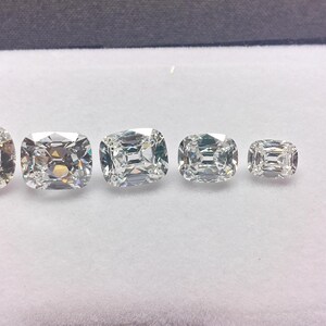 May include: Five cushion-cut diamonds of different sizes are shown on a white surface. The diamonds are clear and faceted, reflecting light. The largest diamond is on the left, with sizes decreasing to the right. The diamonds are set against a white background.