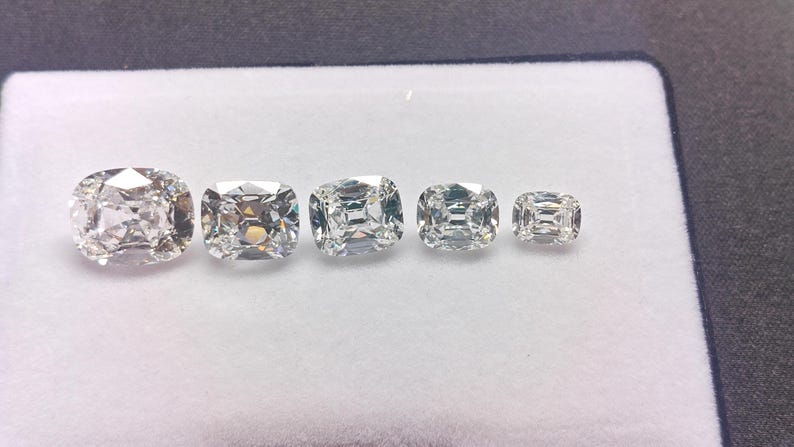May include: Five cushion-cut diamonds of different sizes are shown on a white surface. The diamonds are clear and faceted, reflecting light. The largest diamond is on the left, with sizes decreasing to the right. The diamonds are set against a white background.