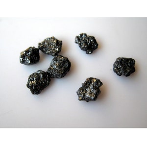Puede incluir: Una colecci&oacute;n de siete diamantes negros, tambi&eacute;n conocidos como diamantes carbonado, sobre una superficie blanca. Los diamantes son en bruto y sin pulir, con un brillo met&aacute;lico oscuro.