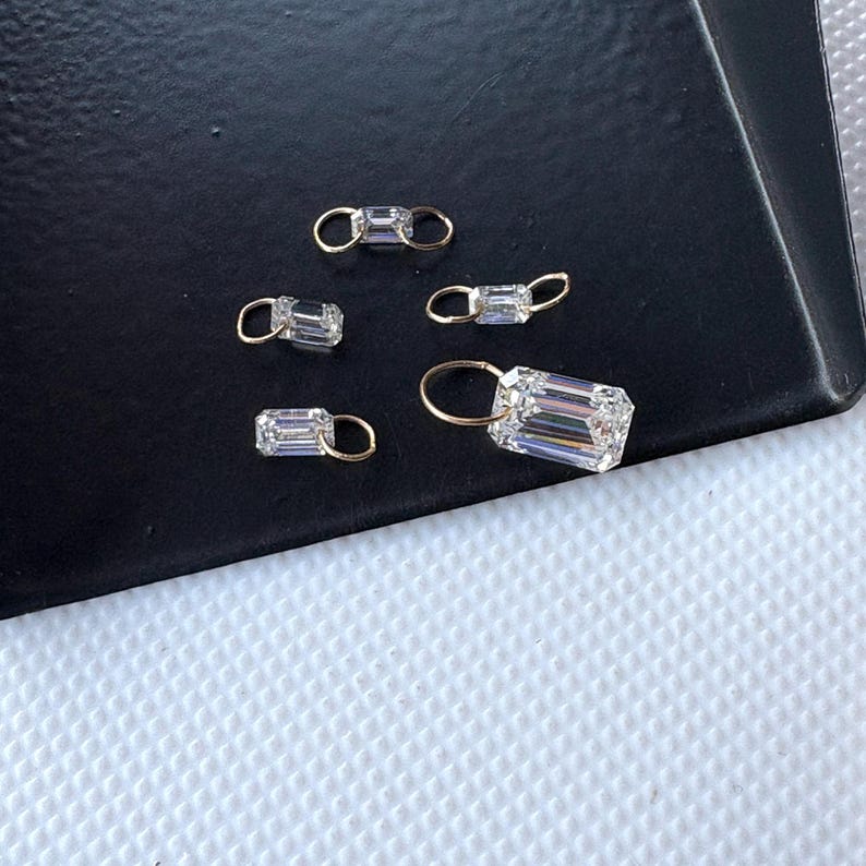 May include: Five small, clear, rectangular-cut crystal charms with gold-colored loops. The charms are arranged on a black surface, with a white textured surface in the background. The crystals reflect light, creating a sparkling effect.