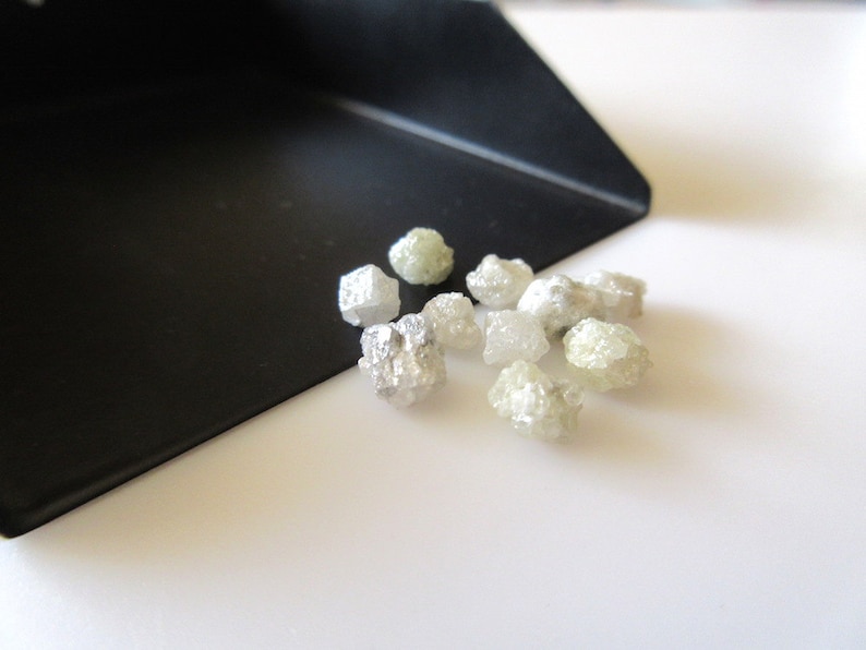 Puede incluir: Una colecci&oacute;n de peque&ntilde;os diamantes en bruto, blancos y verdes. Los diamantes est&aacute;n esparcidos sobre una superficie blanca.