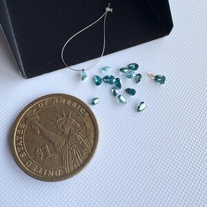 May include: A collection of small, teardrop-shaped, teal-colored gemstones scattered on a white surface. A US one-dollar coin is next to the gemstones for size comparison. A small black tray and a silver wire with a gemstone are also present.