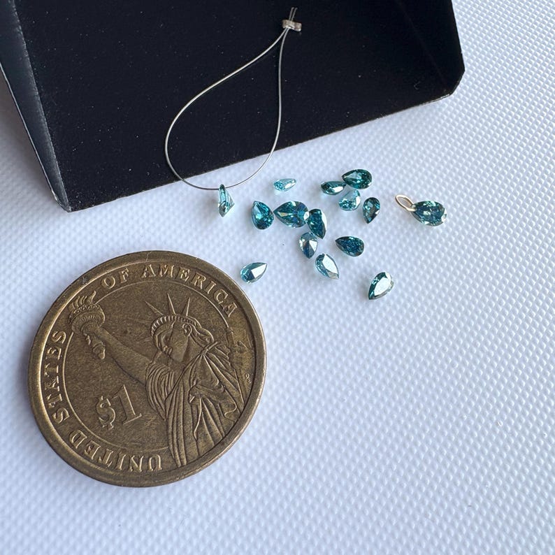 May include: A collection of small, teardrop-shaped, teal-colored gemstones scattered on a white surface. A US one-dollar coin is next to the gemstones for size comparison. A small black tray and a silver wire with a gemstone are also present.