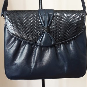 May include: A navy blue leather shoulder bag with a snakeskin flap and a bow detail. The bag has a single strap and a flap closure.