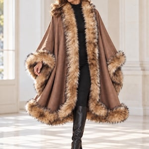 May include: A brown suede-textured cape featuring a thick, fluffy faux fur trim along the front opening and the hemline. The garment has wide, open arm slits and a loose, flowing silhouette that drapes over a black turtleneck and black boots.