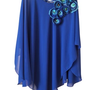 May include: A royal blue cape top with a round neckline and a floral shoulder detail. The flowers are in shades of blue and embellished with sequins. The fabric is sheer and has a flowing design. The top is on a black hanger.