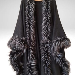 May include: A black cape with faux fur trim. The cape has a wide collar and trim along the edges and sleeves. The faux fur is a mix of black and grey, creating a striking contrast against the solid black fabric.