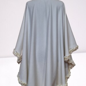 Back view of an off-white cashmere bridal cape with luxurious textured trim outlining the hem and neckline. Lightweight, flowing winter wedding shawl with a timeless vintage-inspired design.