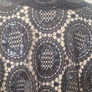 May include: Close-up of a black lace top with a high neckline. The lace features a circular, openwork design with sequins. The top is worn over a neutral-colored underlayer, creating a contrast. The garment appears to be a formal or evening wear piece.