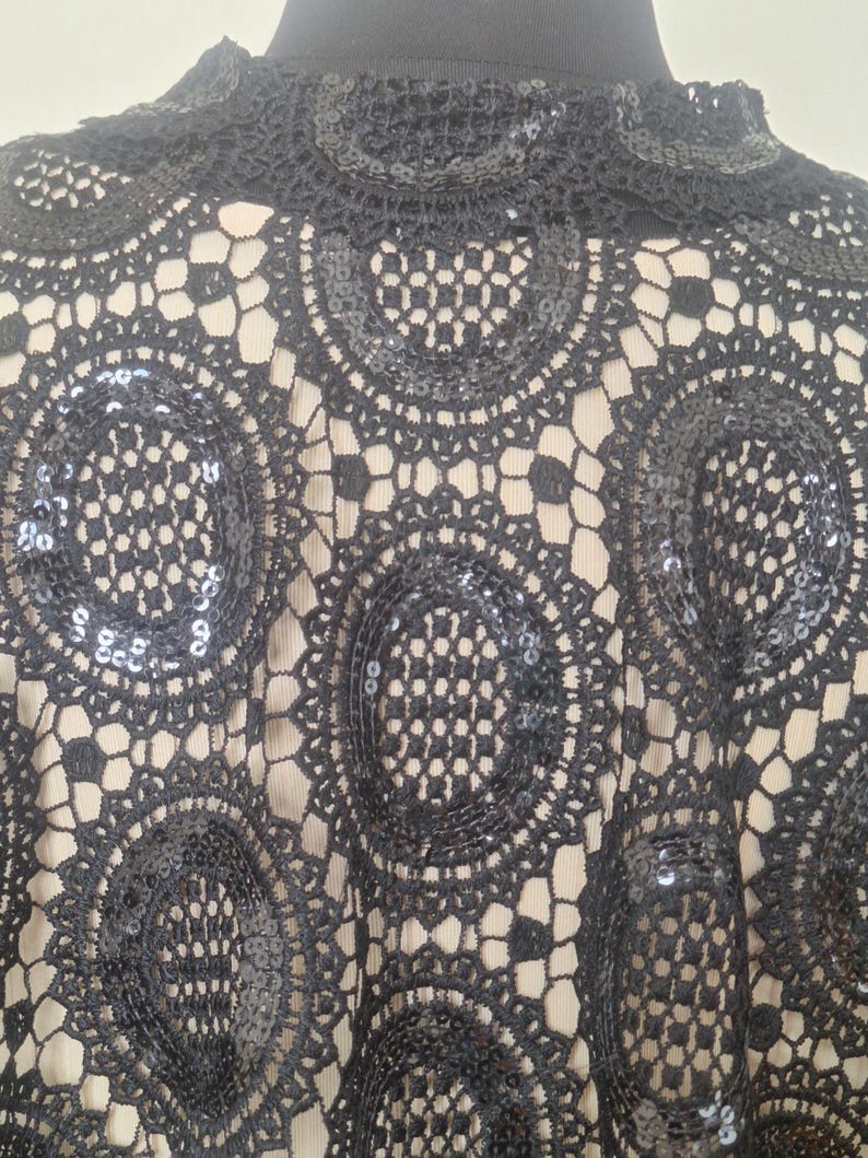 May include: Close-up of a black lace top with a high neckline. The lace features a circular, openwork design with sequins. The top is worn over a neutral-colored underlayer, creating a contrast. The garment appears to be a formal or evening wear piece.