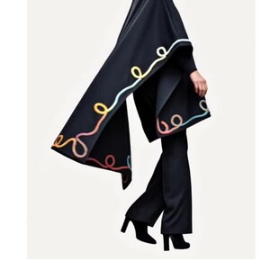 A black cape featuring a decorative trim of swirling, colorful yarn in shades of light blue, yellow, and peach. The cape is worn over a black turtleneck sweater, black trousers, and black ankle boots, creating a monochromatic ensemble.