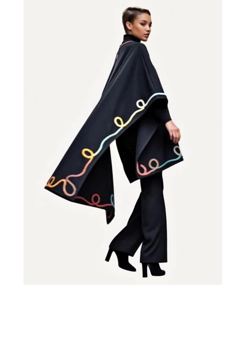 A black cape featuring a decorative trim of swirling, colorful yarn in shades of light blue, yellow, and peach. The cape is worn over a black turtleneck sweater, black trousers, and black ankle boots, creating a monochromatic ensemble.
