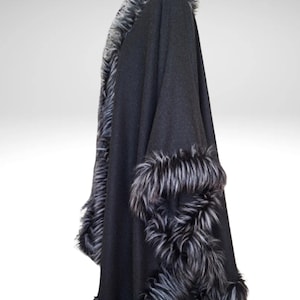 May include: A black cape with faux fur trim. The cape is made of a dark fabric and features a thick, fluffy trim in shades of grey and white along the edges and cuffs. The garment is draped, showcasing its shape.