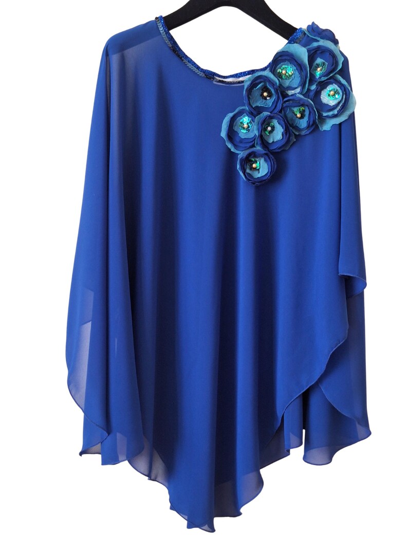 May include: A royal blue cape top with a round neckline and a floral shoulder detail. The flowers are in shades of blue and embellished with sequins. The fabric is sheer and has a flowing design. The top is on a black hanger.