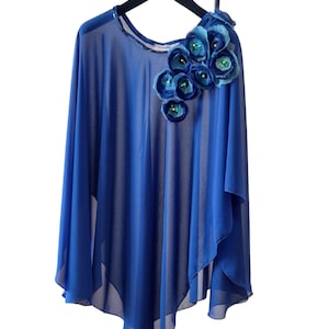 May include: A flowing, cobalt blue cape-style top with a decorative floral embellishment. The flowers are in shades of blue and teal, with sequin centers. The top is sheer and hangs from a black hanger.