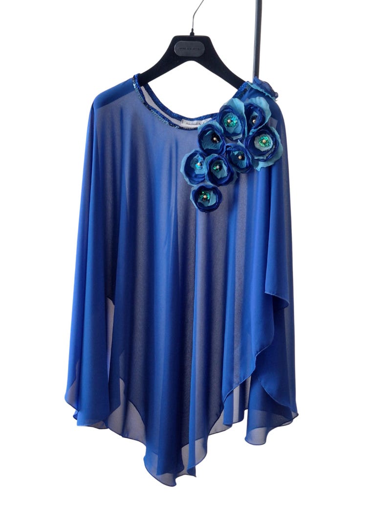 May include: A flowing, cobalt blue cape-style top with a decorative floral embellishment. The flowers are in shades of blue and teal, with sequin centers. The top is sheer and hangs from a black hanger.