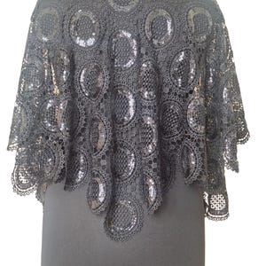 May include: Black lace cape with a high collar. The cape features a circular pattern with sequin embellishments. The garment is displayed on a black mannequin, showcasing its design and texture. The cape is a formal wear accessory.