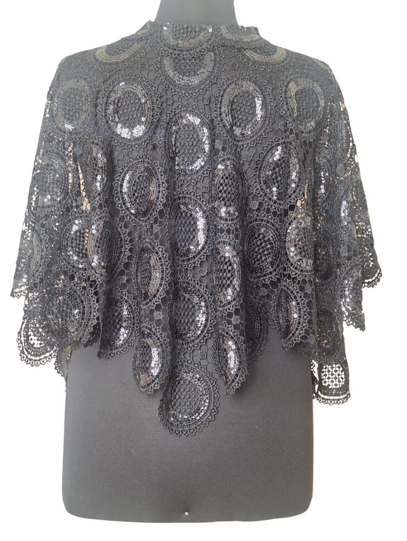 May include: Black lace cape with a high collar. The cape features a circular pattern with sequin embellishments. The garment is displayed on a black mannequin, showcasing its design and texture. The cape is a formal wear accessory.
