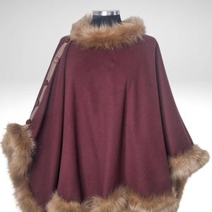May include: A burgundy cape with faux fur trim around the collar and hem. The cape has a button closure on the left shoulder and is displayed on a mannequin. The faux fur is a light brown colour.