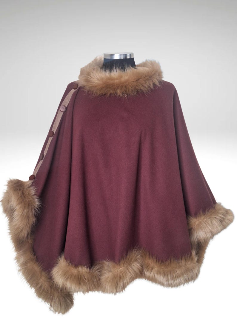 May include: A burgundy cape with faux fur trim around the collar and hem. The cape has a button closure on the left shoulder and is displayed on a mannequin. The faux fur is a light brown colour.