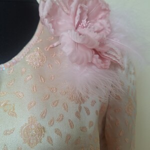 May include: A light pink dress with a floral design and a pink flower with feathers on the shoulder.