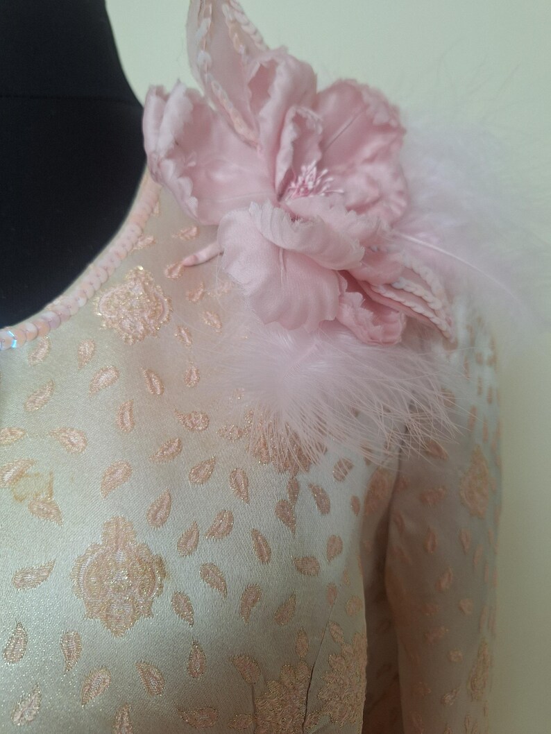 May include: A light pink dress with a floral design and a pink flower with feathers on the shoulder.