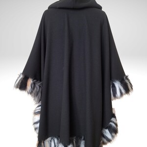 May include: A black hooded cape with faux fur trim. The cape features a hood and is trimmed with black, white, and tan faux fur along the edges of the sleeves and bottom hem. The cape is made of a soft, flowing fabric.