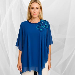 May include: A cobalt blue, flowing top with a decorative floral embellishment on the shoulder. The top features a round neckline with a beaded trim and short, wide sleeves. The model is wearing cream-coloured trousers.