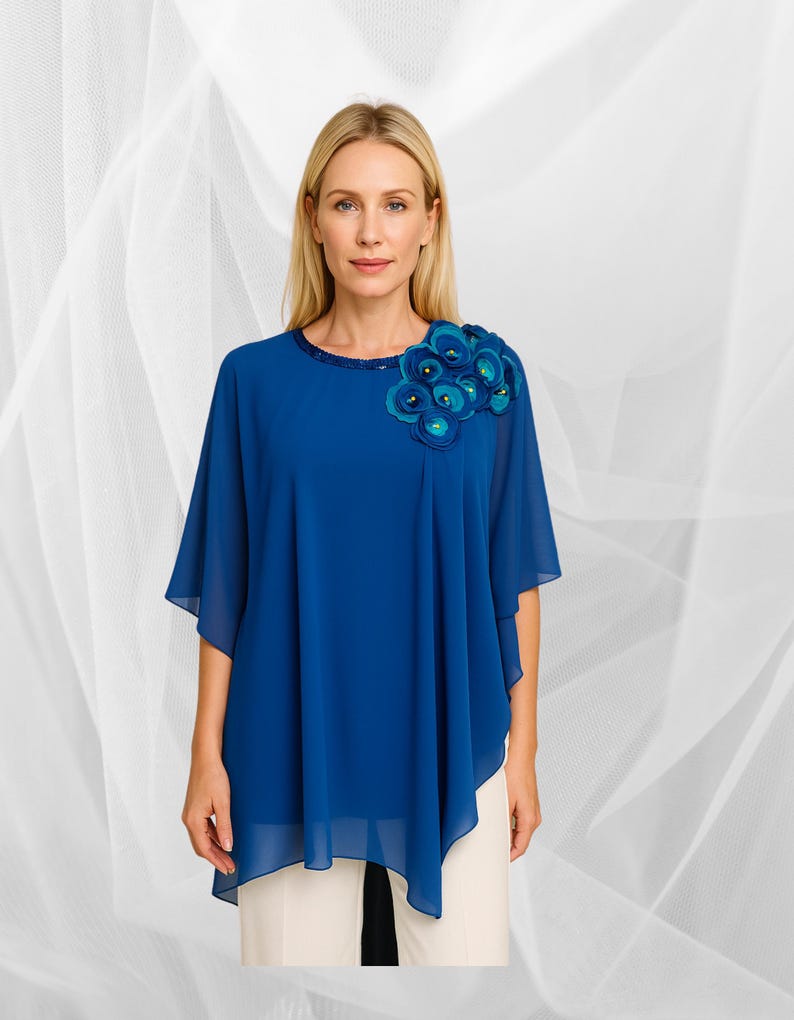 May include: A cobalt blue, flowing top with a decorative floral embellishment on the shoulder. The top features a round neckline with a beaded trim and short, wide sleeves. The model is wearing cream-coloured trousers.