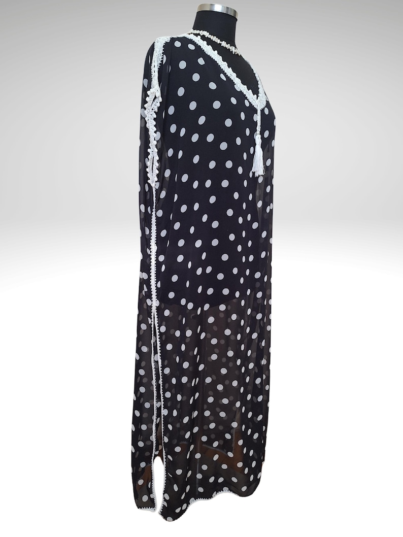 May include: A black and white polka dot dress with white lace trim and a tassel. The dress is displayed on a mannequin. The dress has a V-neckline and long sleeves. The dress is made of a lightweight fabric.