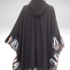 May include: A black hooded cape with faux fur trim. The cape features a hood and is trimmed with a mix of black, white, and tan faux fur along the edges and hood. The cape is displayed against a white background.