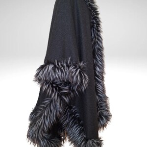May include: Black cape with faux fur trim. The cape has a hood and is trimmed with long, grey and black faux fur along the edges and hood. The cape is made of a soft, dark fabric.