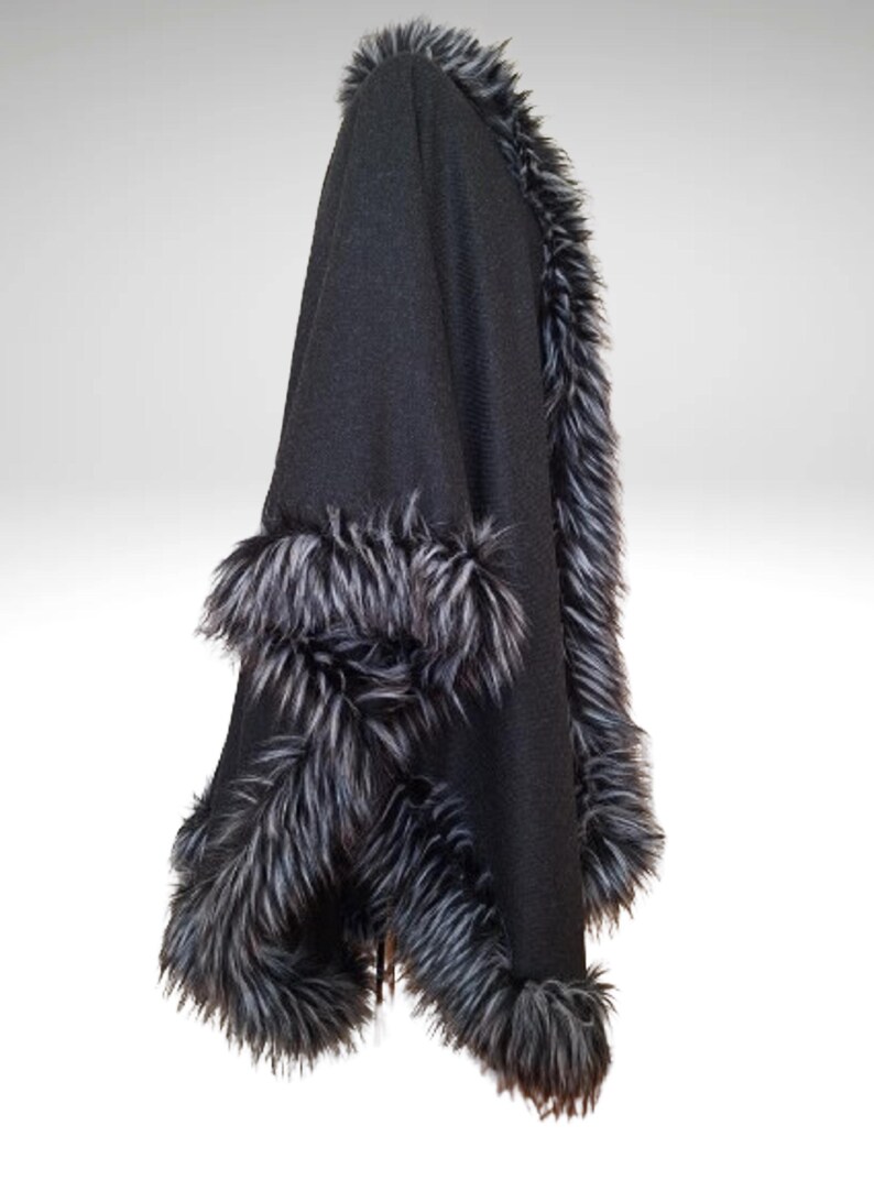 May include: Black cape with faux fur trim. The cape has a hood and is trimmed with long, grey and black faux fur along the edges and hood. The cape is made of a soft, dark fabric.