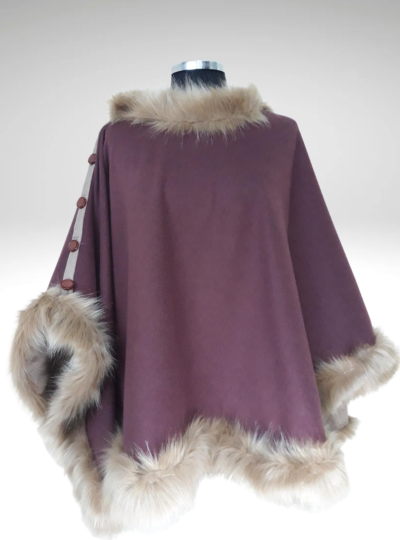 May include: A plum-coloured cape with a faux fur collar and trim. The cape features decorative buttons on the sleeve and is displayed on a mannequin. The fur trim is a light brown colour.