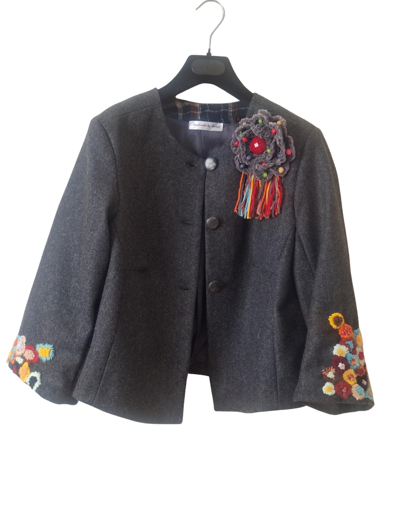 A gray wool cropped jacket with a Handmade Detachable Brooch and colorful hand embroidery on the cuffs. The jacket has a button closures and is hanging on a black hanger.