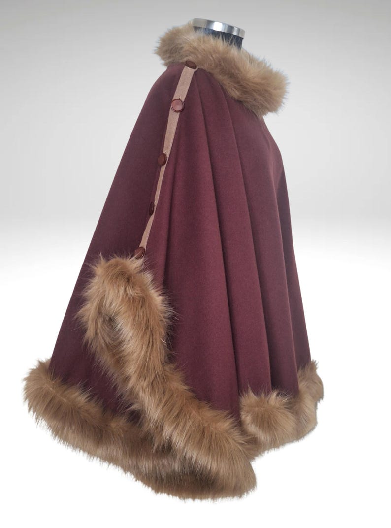 May include: A burgundy cape with faux fur trim and collar. The cape has a tan accent strip with brown buttons. The faux fur is light brown. The cape is a winter garment.