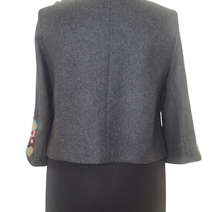 May include: A gray wool jacket with colorful embroidered details on the sleeves. The jacket has a round neckline and a cropped length.