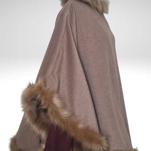 May include: A brown cape with faux fur trim around the collar and edges. The cape is made of a soft material and has a burgundy accent on the inside. The faux fur is a light brown colour.